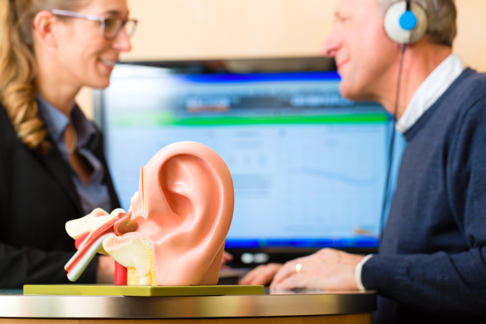 A clinician and a patient wearing headphones during a hearing test, with an ear model in the foreground
